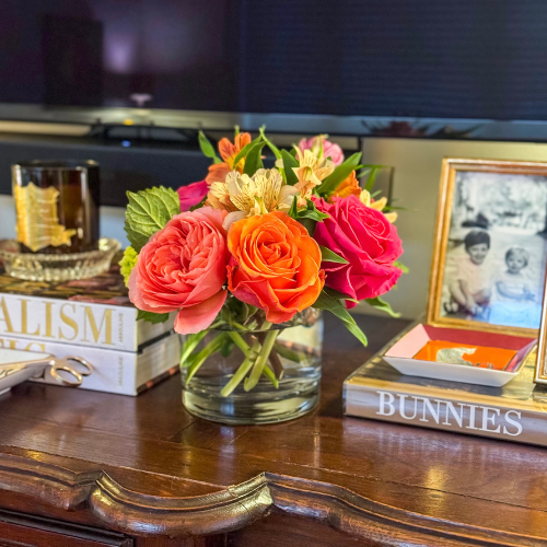 Bouquet of colorful flowers in a clear vase on a wooden surface with books and a photo frame.