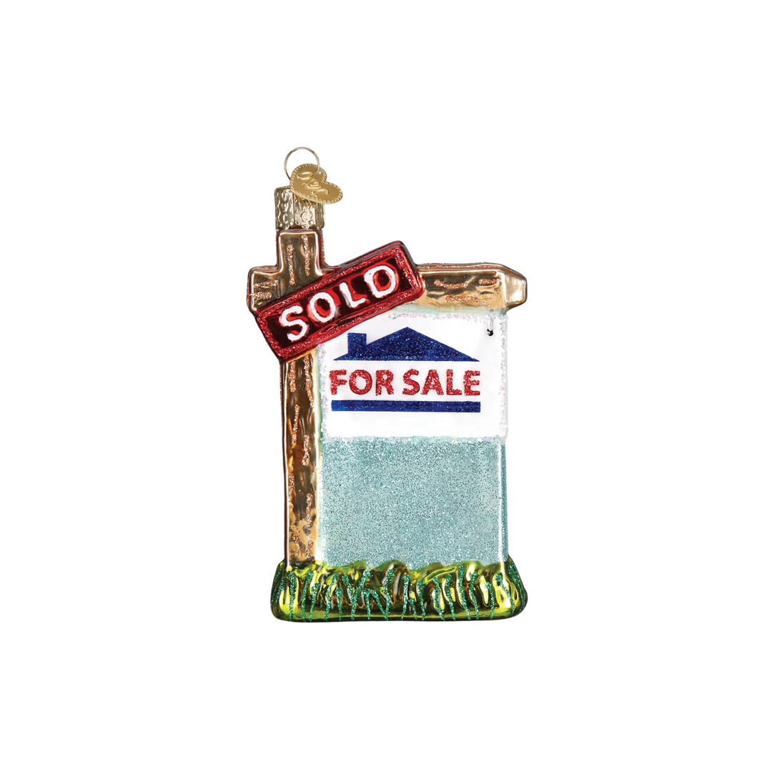 Realty Sign Ornament by Old World Christmas — red and white “SOLD” glass ornament hanging on a Christmas tree.