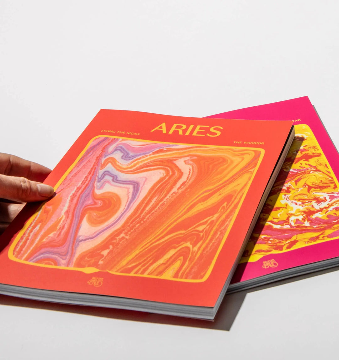 Hand holding the Aries: The Warrior astrology workbook by In Case of Emergence over a pink workbook