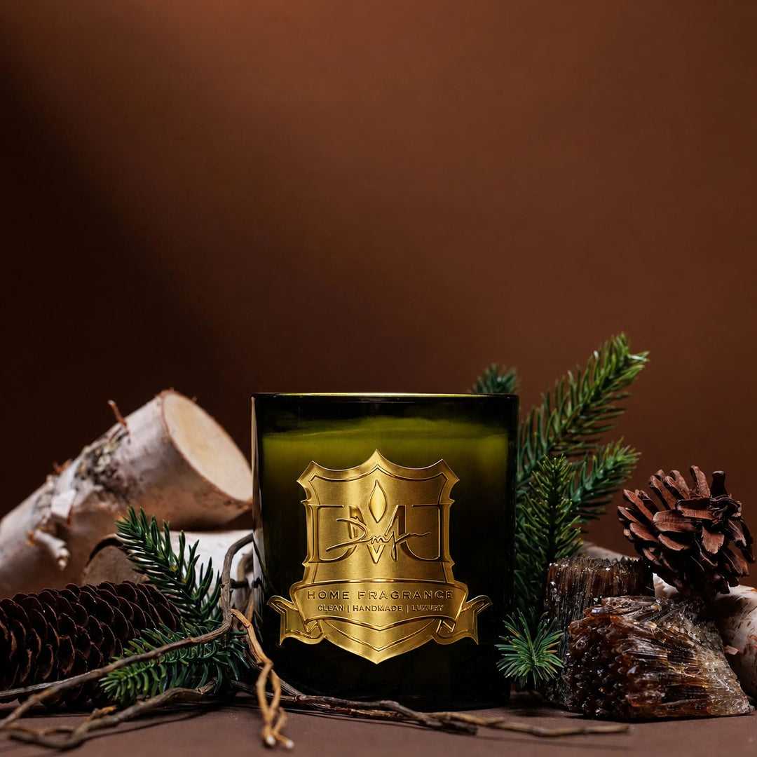 Woods Candle by DMJ Home Fragrance, olive green glass with gold crest label, styled with spruce branches, birch logs, pinecones and amber resin on a warm brown backdrop