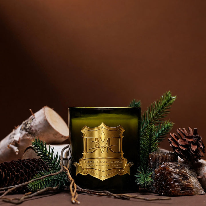 Woods Candle by DMJ Home Fragrance, olive green glass with gold crest label, styled with spruce branches, birch logs, pinecones and amber resin on a warm brown backdrop