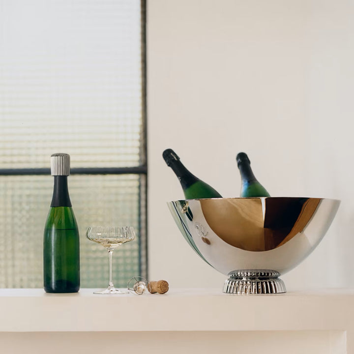 Georg Jensen Bernadotte champagne stopper on a bottle beside a stainless steel champagne cooler and coupe glass
