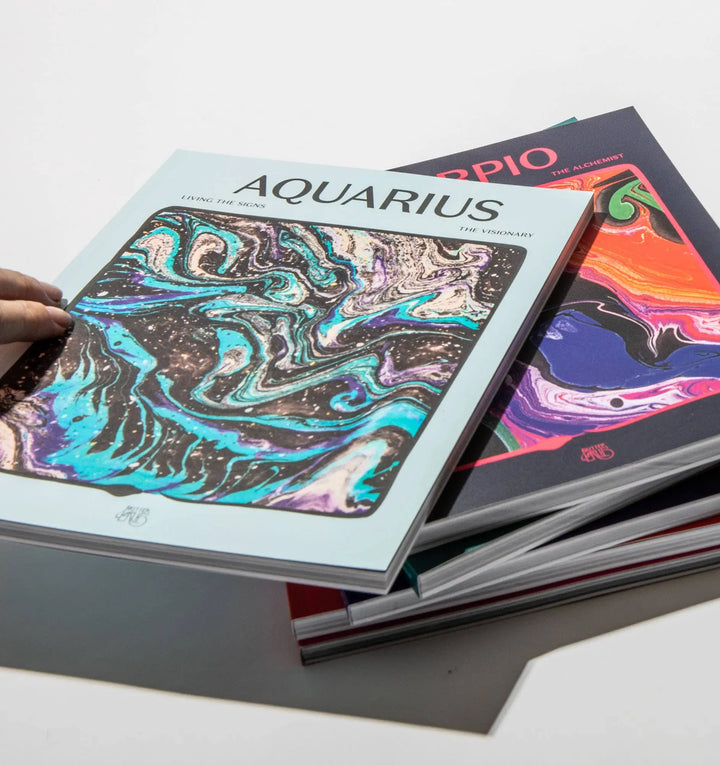 Hand holding Living the Signs: Aquarius, The Visionary above a stack of other In Case of Emergence zodiac workbooks
