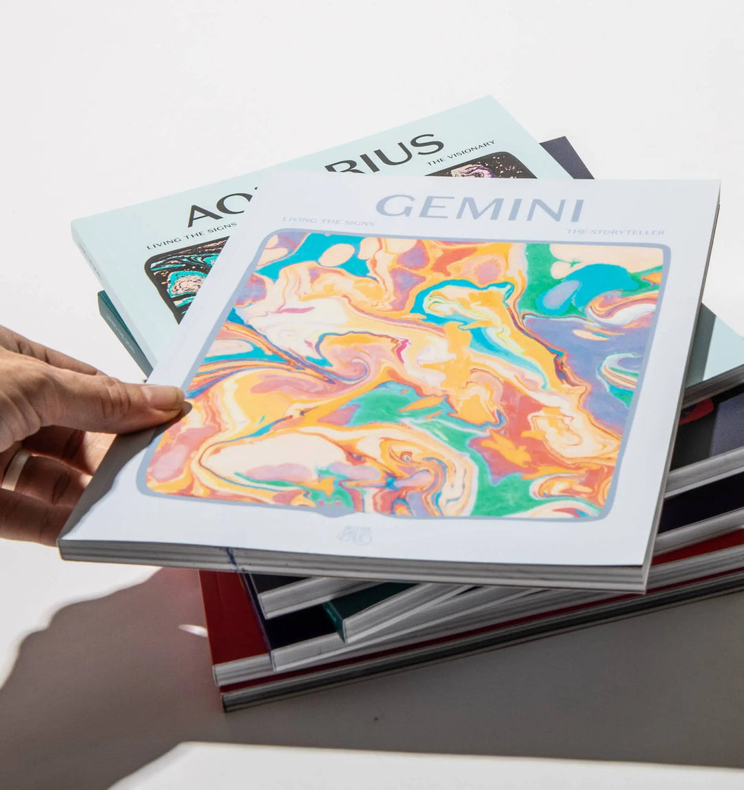 Hand holding Living the Signs: Gemini over the Aquarius workbook and a stack of the zodiac series
