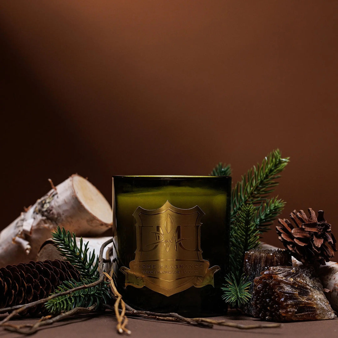 Woods luxury Christmas candle by DMJ Home Fragrance in signature glass vessel surrounded by pine branches, birch logs, and pinecones — hand-poured coconut wax holiday candle with white pine, fir balsam, birchwood, musk, and amber.